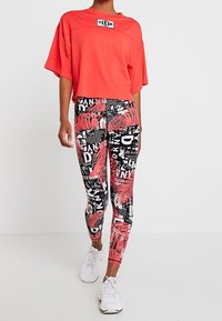 Röd kort t-shirt med logotyp; svarta leggings med ett djärvt, färgglatt palmmönster och text i vitt, rosa och svart. Vita sneakers.