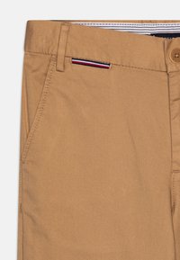 Chinos in cotone marrone con texture a spina di pesce diagonale, dotati di una tasca frontale e un accento di cucitura a contrasto in rosso, bianco e blu.