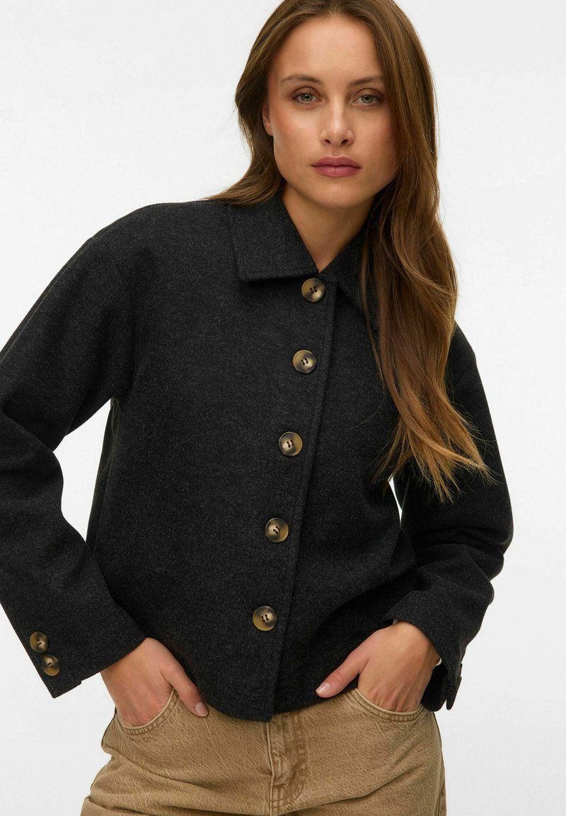 Femme aux cheveux longs châtains clairs portant une veste boutonnée gris foncé et un pantalon beige, debout les mains dans les poches sur un fond blanc.