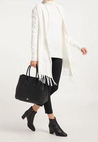 Sac en cuir noir avec deux poignées, surface texturée et accent logo. Associé à un châle blanc, un col roulé et des bottines noires.