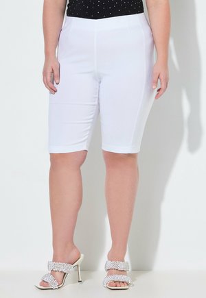 Žena v bílých šortkách sahajících ke kolenům, černém topu s bílými puntíky a bílých sandálech na podpatku s ozdobnými korálky.