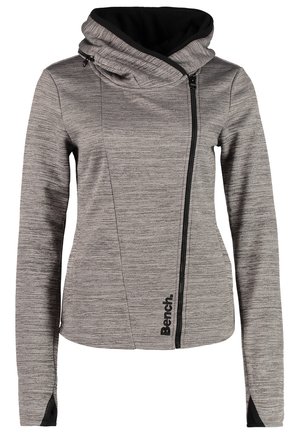 Frau trägt einen grauen Hoodie mit Reißverschluss und schwarzer Kapuze, blaue Skinny-Jeans, weißes Oberteil und dunkelblaue Sneaker mit weißen Schnürsenkeln.