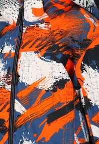 Abstrakt orange, marinblå, vit och svart färgstänkmönster på en jacka med dragkedja framtill, med en liten "CMP"-logotyp på axeln.