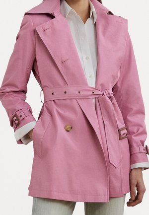 Mujer con gabardina rosa con cinturón sobre una camisa blanca abotonada, con una mano en el bolsillo, mostrada desde los hombros hasta los muslos.