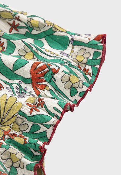 Tissu blanc avec un motif floral vert, jaune, rouge et orange, comportant un bord froncé avec une couture rouge sur un fond gris clair.