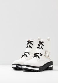 Bottes blanches à la cheville en cuir embossé. Dotées de lacets noirs, d'une boucle latérale et d'une semelle épaisse noire avec un accent métallique.