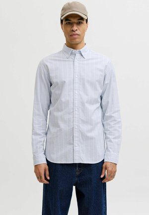 Mies, jolla on vaaleansininen raidallinen nappipaita, tummansiniset farkut ja beige lakki, seisomassa valkoista taustaa vasten.