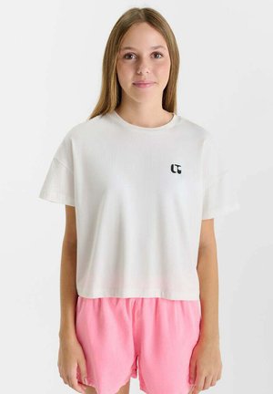 Jeune femme aux cheveux châtains clairs et raides, portant un t-shirt court blanc avec un logo et un short rose, debout devant un fond blanc uni.