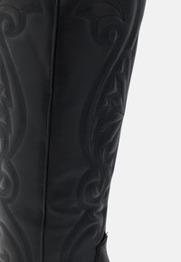 Bota de cuero negro hasta la rodilla con intrincado bordado tonal y elementos de diseño curvados a lo largo del eje, con una textura suave.