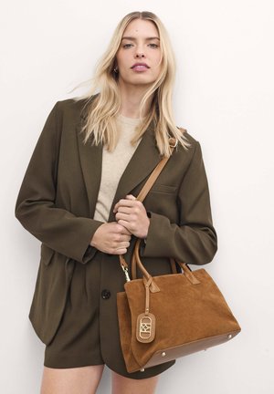 Brown suede handbag with leather accents, rectangular shape, short handles, and a gold hardware tag. Model wears an olive blazer and beige top.
