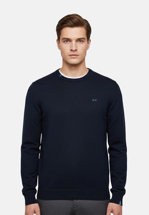 Giovane uomo con capelli castani corti che indossa un maglione girocollo blu navy semplice con un piccolo logo azzurro sul petto e pantaloni grigi.