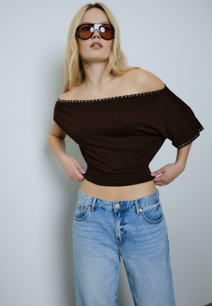 Donna bionda che indossa una maglia marrone oversize con spalle scoperte e occhielli, jeans azzurri chiari e grandi occhiali da sole marroni contro uno sfondo semplice.