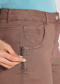 Hände ziehen den kleinen Reißverschluss der rechten vorderen Tasche von braunen Hosen, die mit einem türkisfarbenen Oberteil kombiniert sind.