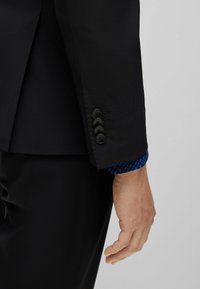 Manica di giacca nera con quattro bottoni neri e polsino di camicia blu testurizzato con un motivo geometrico, mano visibile riposante di lato.
