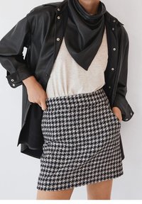 Femme portant une veste en cuir noire, une écharpe noire, un haut blanc et une jupe pied-de-poule noire et blanche, les mains dans les poches.