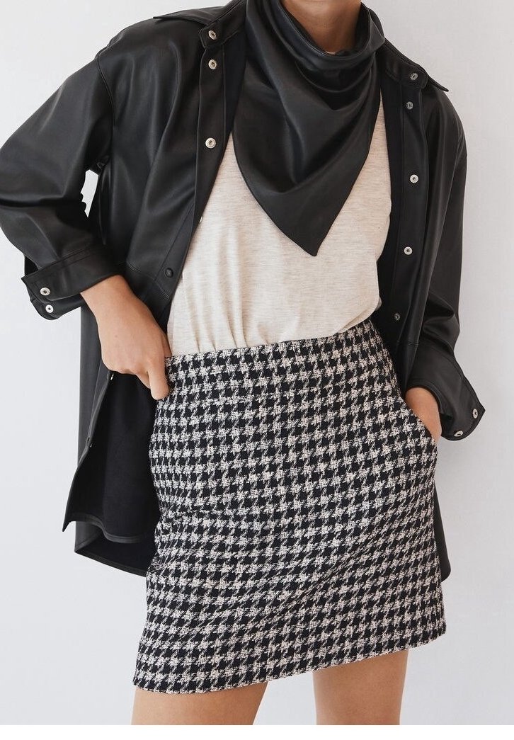 Femme portant une veste en cuir noire, une écharpe noire, un haut blanc et une jupe pied-de-poule noire et blanche, les mains dans les poches.