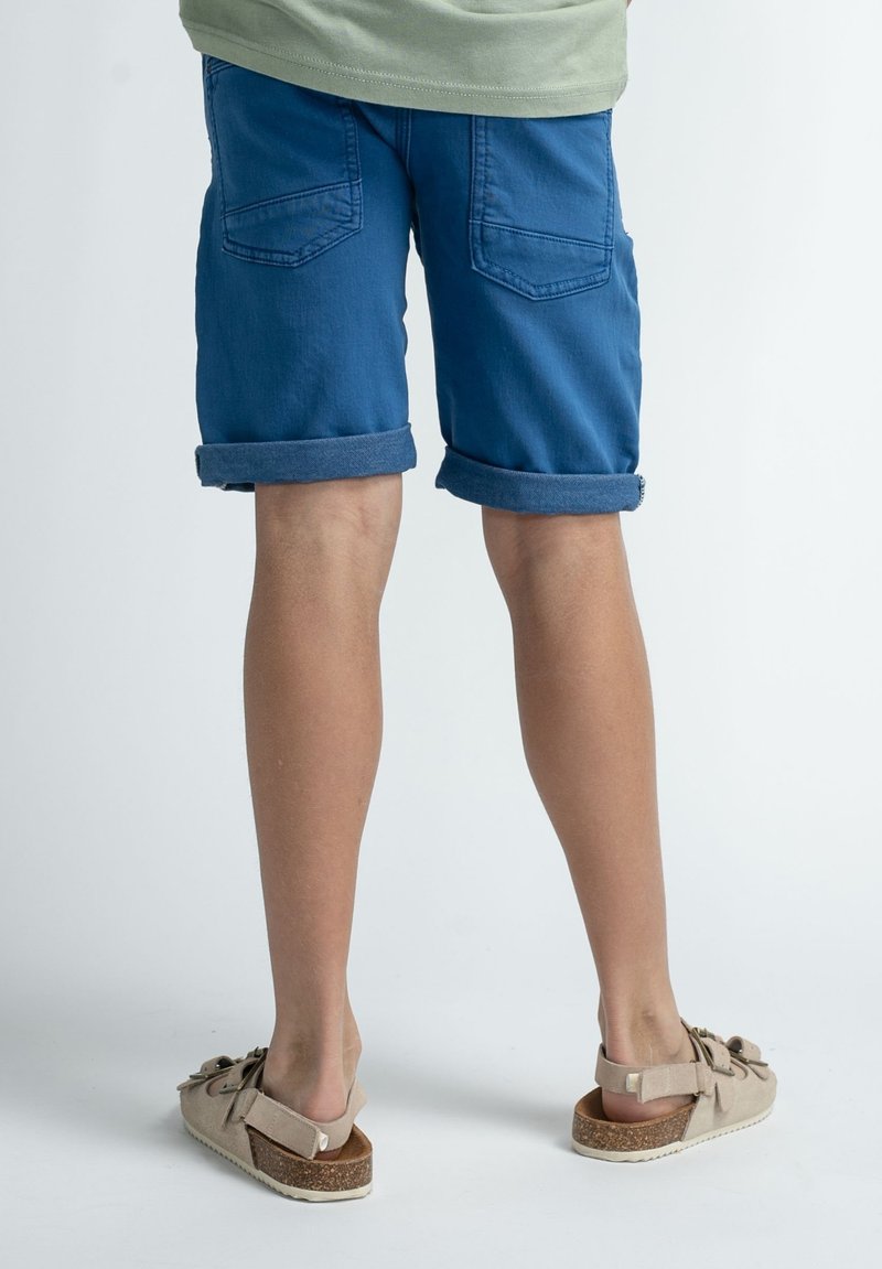 Pantalones cortos de mezclilla azul con dobladillos enrollados, que cuentan con dos bolsillos traseros, combinados con sandalias de color beige claro con suela de corcho y acentos de lazo.