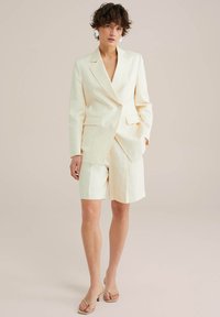 Světle žlutý přizpůsobený blazer s jediným knoflíkem, spárovaný s odpovídajícími šortkami ke kolenům. Texturovaná tkanina s předními kapsami.