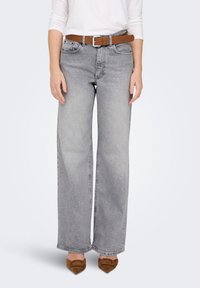 Graue Jeans mit weitem Bein und gesprenkeltem Muster, ausgestattet mit vorderen Taschen und einem braunen Leder Gürtel. Das Modell trägt braune Absatzschuhe.