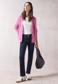 Cardigan de punto rosa sobre una camiseta blanca, combinado con unos jeans oscuros y zapatillas blancas. Sosteniendo un bolso gris, de pie sobre un fondo claro.