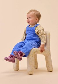 Un tout-petit avec des cheveux blonds portant une salopette bleue et des chaussures roses assis sur une petite chaise tachetée contre un fond beige.