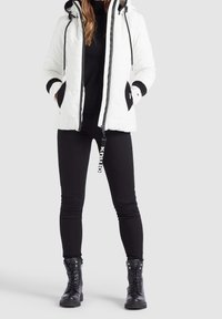 Veste rembourrée blanche avec une capuche, intérieur noir et poignets noirs contrastés. Portée sur un haut noir et associée à un pantalon skinny noir et des bottes.