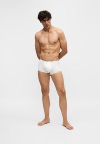 Jeune homme debout pieds nus, portant un boxer blanc, les bras derrière le dos, sur un fond clair uni.