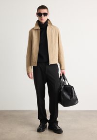 Tan suede jacket with a high collar, paired with a black turtleneck sweater and black trousers, holding a black leather bag. Black sunglasses.