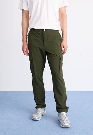 Olive-green cargo pants with multiple side pockets, zipper detail, and a relaxed fit, paired with light gray athletic shoes on a blue floor.
