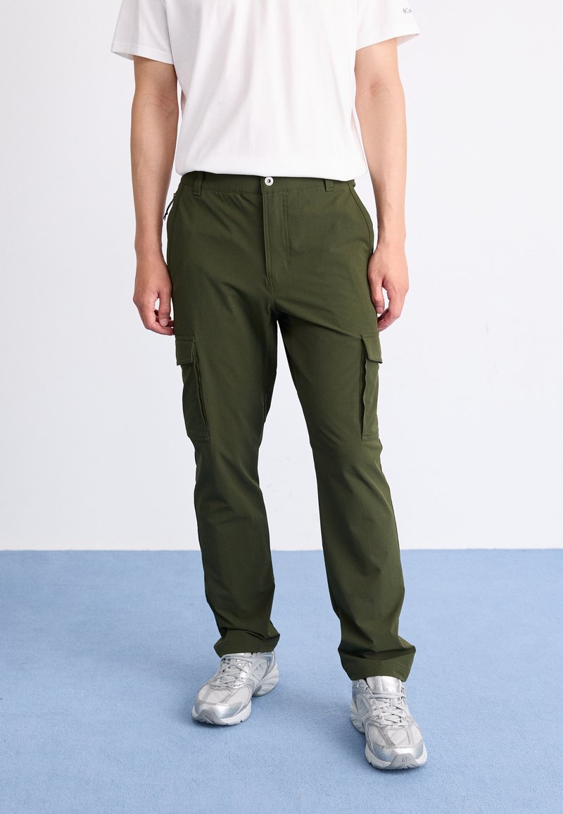 Pantalones cargo verde oliva con múltiples bolsillos laterales, detalles de cremallera y un ajuste relajado, combinados con zapatillas deportivas gris claro sobre un suelo azul.
