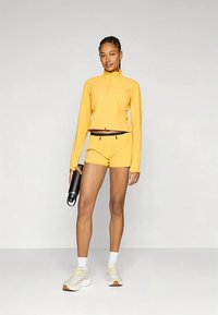 Gelbes langärmeliges Sportoberteil mit Reißverschluss und zwei vorderen Taschen, kombiniert mit passenden Shorts und weißen Turnschuhen, hält eine schwarze Wasserflasche.