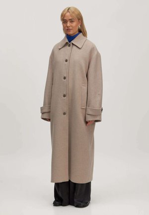 Beige Wollmischungsjacke, mittellang, mit spitzem Kragen und Knopfschluss. Verfügt über seitliche Taschen und weite Ärmel, kombiniert mit schwarzen Hosen.