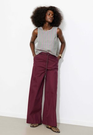 Femme avec une grande coiffure afro se tient contre un mur blanc, portant un débardeur rayé, un pantalon large bordeaux et des sandales.