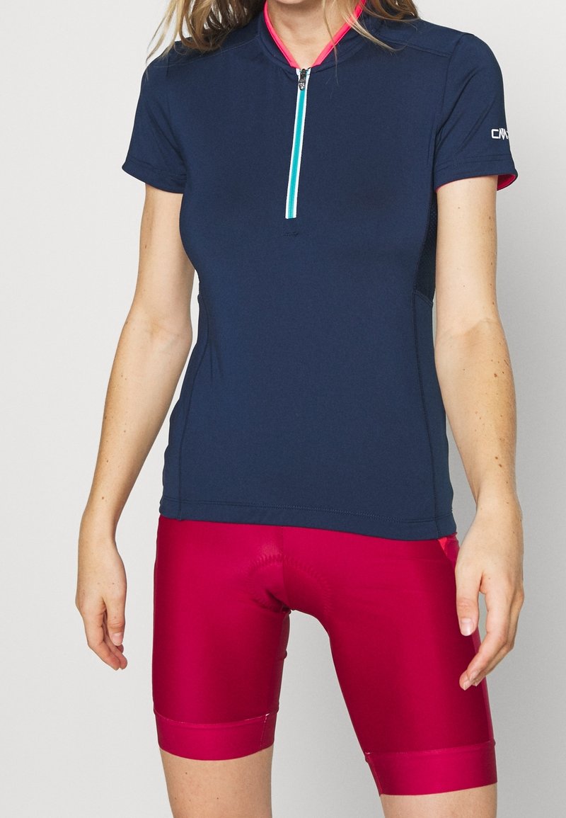 Femme portant un maillot de cyclisme bleu marine à manches courtes avec une fermeture éclair à l'avant et un cuissard de cyclisme rembourré rose vif.