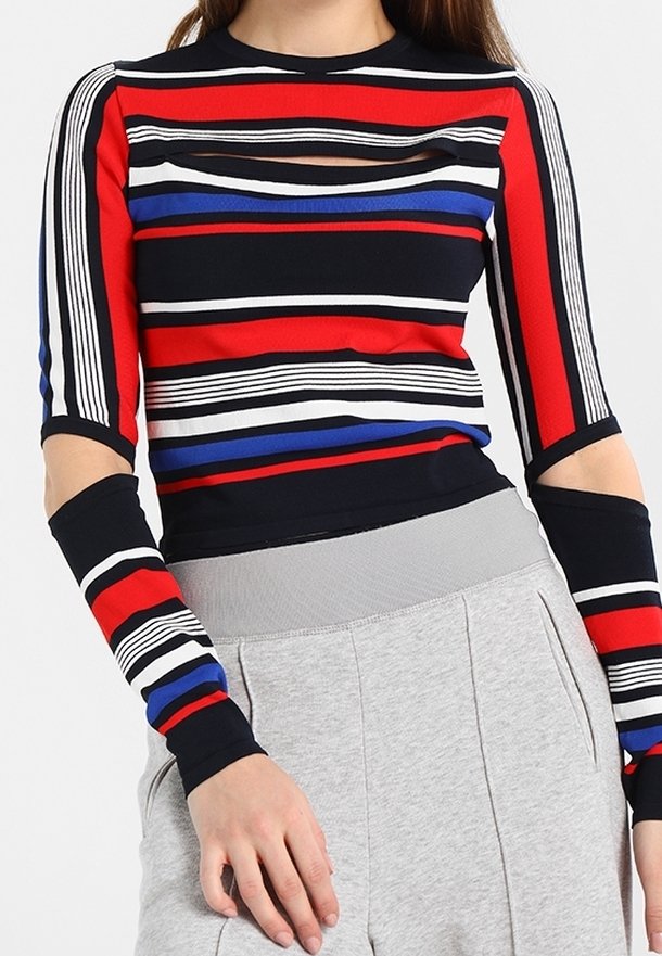 Femme portant un haut à manches longues rayé multicolore avec des découpes sur les manches et la poitrine, associé à un pantalon gris clair.