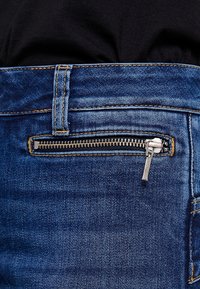 Vaqueros de mezclilla azul con un bolsillo lateral con cremallera y herrajes plateados, textura desgastada y costuras en contraste de color amarillo.