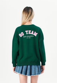 Sweatshirt vert arborant "GO TEAM" en lettres roses, avec "GAP ATHLETICS EST. 1969" en dessous. Porté avec une jupe plissée en denim.
