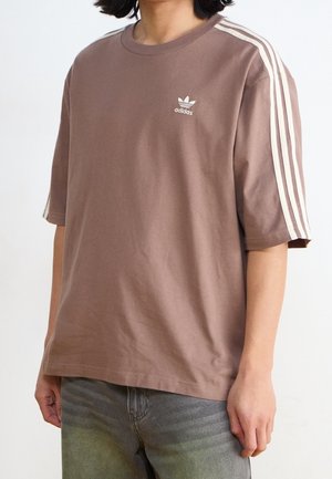 Person som bär en lös brun Adidas-t-shirt med vita ränder på ärmarna och logotypen på bröstet, ihop med blekta grå jeans.