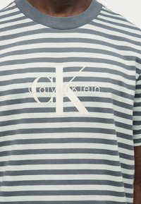 Άνδρας που φοράει γκρι και λευκό T-shirt με οριζόντιες ρίγες και μεγάλο λευκό λογότυπο "CK" και "Calvin Klein" στο στήθος.