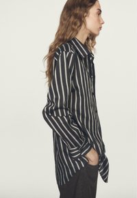 Czarna i biała koszula z długim rękawem w paski, z klasycznym kołnierzykiem i luźnym krojem, noszona na ciemnych spodniach.