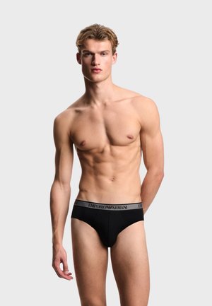 Giovane modello maschile in piedi, indossa slip neri Emporio Armani con fascia con logo, su sfondo grigio chiaro uniforme.