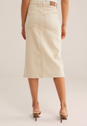 Bež jeans krilo do kolen z zadnjimi žepi, nošeno z belimi sandali s peto na stoječi osebi, obrnjeni stran.