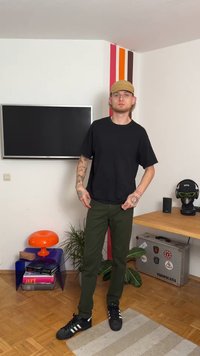 Svart t-skjorte med korte ermer kombinert med olivengrønne bukser og svarte joggesko med hvite striper, plassert i et minimalistisk innendørsmiljø.