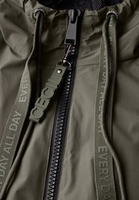 Chaqueta ligera de color verde oliva con cremallera negra, cordón a juego y cuerdas texturizadas con el texto "ICECIL" y "ALL DAY EVERY DAY".