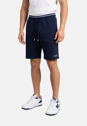 Hombre con pantalones cortos azul marino con cordón y bolsillos laterales, y zapatillas blancas, de pie sobre un fondo blanco liso.