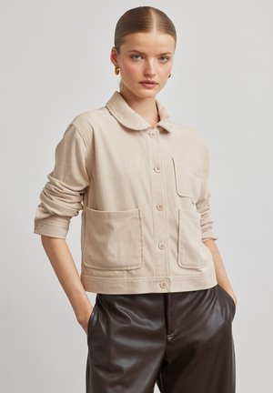 Young woman with hair tied back wearing beige corduroy button-up jacket and dark brown leather pants, standing with hands in pockets.