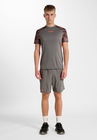 Jonge man staand, gekleed in grijze sportkleding met oranje accenten, zwarte sneakers en witte sokken tegen een effen witte achtergrond.