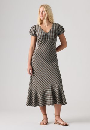 Femme aux longs cheveux blonds portant une robe midi à carreaux marron et bleu avec des manches bouffantes, un col à nouer et un ourlet à volants, chaussée de sandales beiges à lanières.