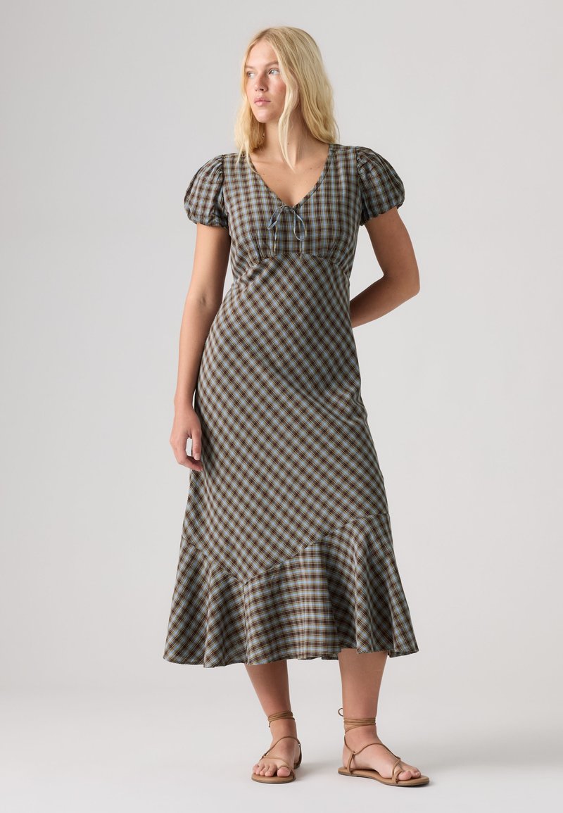 Femme aux longs cheveux blonds portant une robe midi à carreaux marron et bleu avec des manches bouffantes, un col à nouer et un ourlet à volants, chaussée de sandales beiges à lanières.
