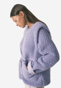 Fioletowa, puszysta kurtka polarowa z ściągaczem w talii i bocznymi kieszeniami. Posiada okrągły dekolt i teksturowany materiał. Akcenty w białym kolorze przy mankietach.